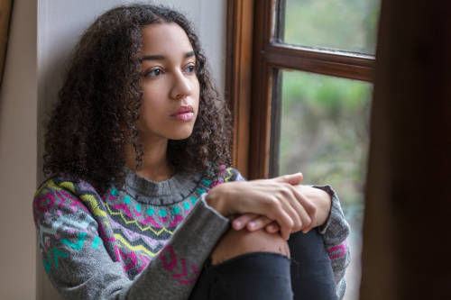 Una adolescente con expresión triste sentada al lado de una ventana