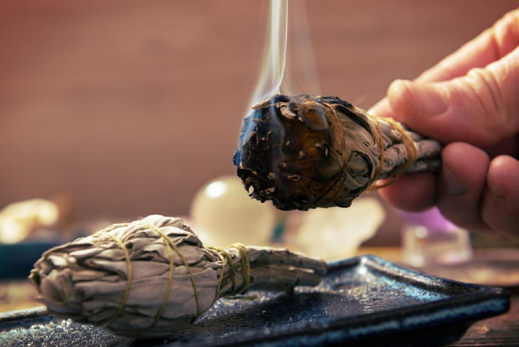 rama de salvia amarrada con un hilo de algodón, esta siendo quemada y desprende humo, esta tomada por una mano y sobre un cenicero