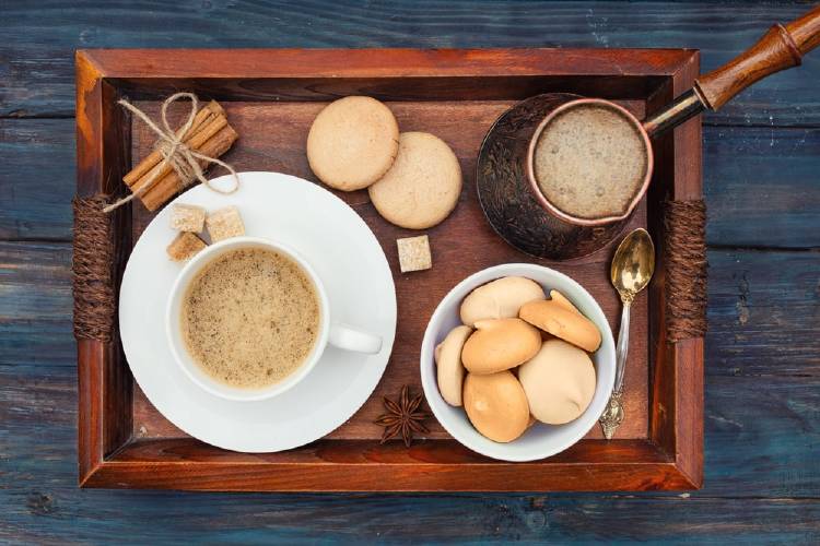 Agrega canela a tu café y tu cuerpo te lo agradecerá. -min bandeja de madera con ramas de canelas amarradas con un lazo, un plato con una taza de café con leche y terrones de azúcar, un bowl con galletas, una cuchara para postres y una jarra de mango largo con cafe con leche. Sobre una mesa de madera azul